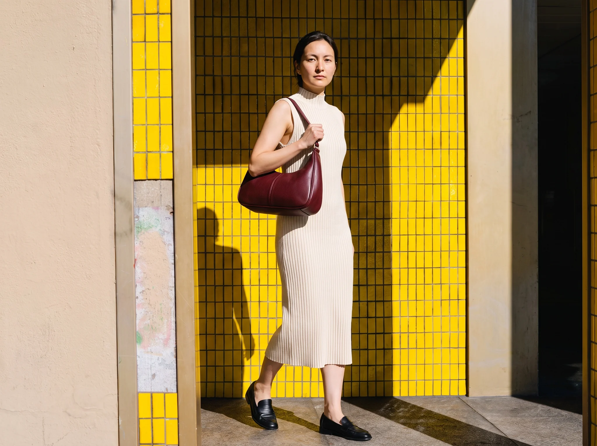 Model carrying a burgundy leather hobo bag by a yellow tiled wall